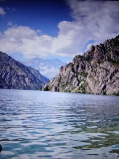 A tranquil lake stretches out towards a dramatic mountainous landscape. The water in the foreground is a mosaic of rippling light blues and whites, suggesting a shallow depth with sunlight reflecting off its surface. Beyond the lake, two imposing mountain ranges rise, their slopes covered in rough, textured rock formations and sparse vegetation. The left mountain is a darker, more rugged mass, while the right mountain, though also rocky, appears slightly less steep and more weathered. In the distance, a hazy range of snow-capped peaks pierces the sky, their outlines softened by atmospheric perspective. The sky above is a vivid blue, dotted with fluffy, white clouds that cast subtle shadows on the mountains. The overall impression is one of serene natural beauty, a secluded mountain environment with a pristine lake at its heart. The image does not depict any people, buildings, or specific urban elements that would place it within the city of Auk, Kyrgyzstan.