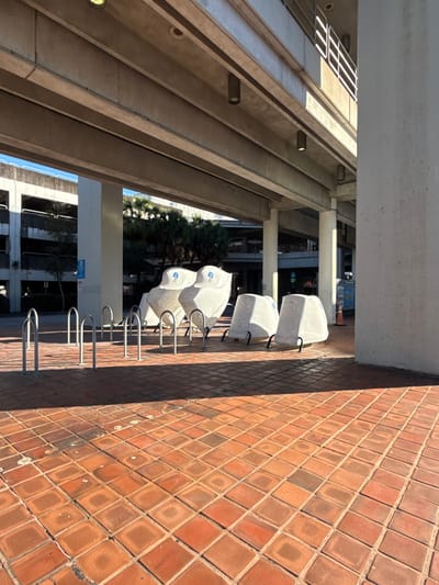 A row of covered stone benches lines a brick walkway under an architectural structure. The benches are white with gray flecks and metal supports. Bike racks are positioned in front of the benches. A multi-story building and trees are visible in the background. The bricks of the walkway are set in a pattern and are various shades of brown.