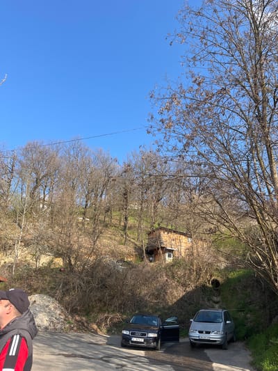 The image shows an outdoor scene in Churka, Bulgaria. Two cars are parked on a narrow paved road: a silver SEAT Alhambra (license plate CN 6365 AH) and a black Audi A4 (license plate K 2676 AT). The Audi's driver's side door is open. The road runs alongside a small wooded area with deciduous trees, mostly bare of leaves. A small wooden house is visible in the trees. A section of a person wearing a black baseball cap and a jacket with a red and white stripe is visible in the lower right corner. The sky is clear and blue, partially obscured by tree branches in the upper left portion of the image. A small pipe or tunnel is located near the cars. The setting appears to be rural or on the edge of a town.