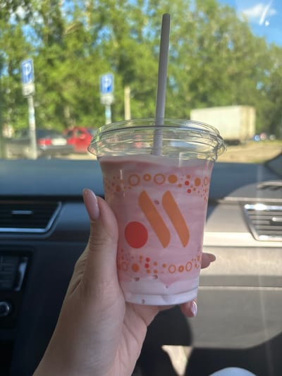 A person's hand with neatly manicured nails holds a plastic cup filled with a pink, frothy beverage and a white straw. The cup has a clear plastic lid and a logo with orange accents and circles. The background is blurred, showing trees, a blue sky, and signs that appear to be parking signs. In the distance, there are vehicles and a building that resembles a truck. The scene appears to be within a car, with the interior of the vehicle visible in the foreground. The image was taken on a sunny day. The location is identified as Fedorino, Russia.