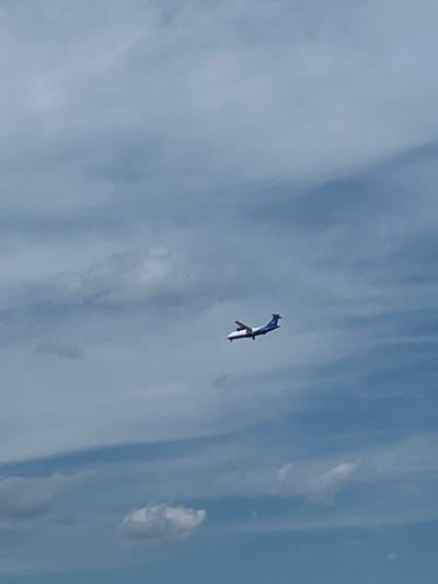 An airplane flies through a cloudy sky. The airplane is white with a blue stripe along the top of its fuselage and tail. It is angled to the left, with its nose pointing slightly upwards. The sky is a pale blue with scattered white clouds of varying densities. The overall impression is one of a clear day with some atmospheric haze. There are no people or other identifiable objects visible in the image. The location of Емельяновское озеро, Russia is not visually indicated in the image.