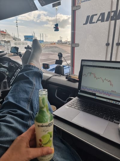 In the cab of a truck, a person's legs are propped up on the dashboard. They are wearing grey socks and blue jeans. In their left hand, they are holding a bottle of "Mojito" flavored drink. To their right, a laptop is open and displaying a financial trading chart. Outside the truck's windshield, a view of a port with a large ship and a blue building is visible. The sky is cloudy. The location is the city of Portsmouth, United Kingdom, specifically within the port area.