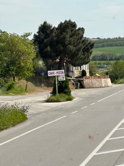 The image shows a roadside scene, likely in a rural area of Λύρα, Greece. A cafe/pizza restaurant is situated near the road, indicated by a sign reading "Cafe Pizza". The building appears to be a multi-story structure with a stone facade and balconies. A large, dark green tree dominates the foreground, partially obscuring the view of the building. The road is paved and features white lane markings. Green vegetation, including trees and shrubs, lines the roadside. The background shows a landscape of rolling hills and fields.