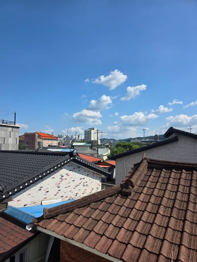 The image captures a sun-drenched, clear blue sky dotted with fluffy white clouds, viewed from an elevated perspective. In the foreground, various rooftops dominate the frame, showcasing different architectural styles and materials. Closest to the viewer are dark, modern-looking tiles, followed by a section of roof with patterned brick-like tiles in red, white, and black. Further in the distance, traditional brown-tiled roofs are visible, some appearing weathered.
Beyond the rooftops, a cityscape unfolds under the bright sky. Buildings of varying heights and designs form a dense urban landscape, with taller structures appearing further back. Glimpses of green trees suggest the presence of natural elements within or surrounding the urban area. The overall scene is one of a pleasant day, likely late morning or early afternoon, given the strong sunlight casting shadows. There are no people or specific activities depicted, making the image a still observation of an urban environment under a clear sky. The orientation of the image is unusual, with the sky occupying the left side and the ground elements on the right, suggesting the photo was taken looking up or tilted. The context of Jincheon-eup, South Korea, points to a specific regional urban setting.