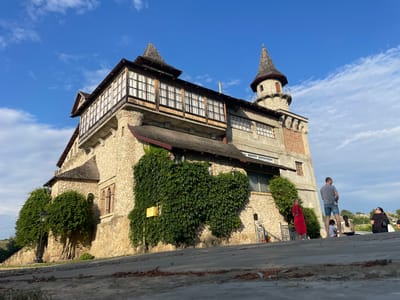 The image depicts a stone-built structure resembling a castle or tower, situated in Colibași, Moldova. The building features multiple levels, a prominent conical turret, and a long, glass-enclosed balcony. The walls are constructed of rough-hewn stones, with some sections covered in dense green ivy. The roof appears to be tiled with a dark material. Several windows are visible, some with visible panes and others in arched openings. The building is set against a backdrop of a bright blue sky with scattered white clouds.
In the foreground, a paved area or walkway is visible, with some debris scattered on it. To the right of the building, a group of people are present. One man, wearing a grey t-shirt and denim shorts, stands facing away from the camera. A woman in a red dress stands near a staircase. Further back, several other individuals are seated or standing, seemingly observing the building or the surroundings. The overall scene suggests a tourist or recreational area.
