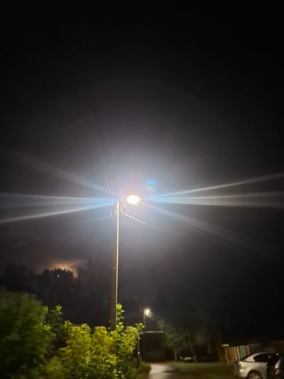 The image depicts a nighttime street scene with a bright streetlamp dominating the center. The streetlamp is tall and metallic, with a single light source emitting a strong, halo-like glow that creates visible light rays fanning out into the darkness. There are power lines attached to the pole. In the background, the sky is dark and overcast, with a faint, hazy illumination suggesting either moonlight through clouds or distant artificial light. Below the streetlamp, trees are visible, some with green foliage, illuminated by the streetlight. To the left, a patch of sky shows a more distinct, diffuse light source, possibly a moon obscured by clouds. On the right side of the image, a residential area is suggested with the outlines of buildings and two parked cars. One car, a silver sedan, is partially visible. The ground appears to be a paved surface, possibly a road or driveway, and there are hints of vegetation along the edges. The scene appears to be an urban or suburban residential area in the city of Novlyanka, Russia. There are no people or animals visible in the image.