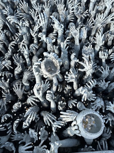 The image displays a dense array of sculptural hands emerging from dark ground, located in Ban Rong Khun, Thailand, at the Wat Rong Khun (White Temple). Hundreds of greyish-white, skeletal-like hands are depicted, reaching upwards from the dark surface. Many hands are posed with open palms or grasping gestures. Two prominent decorative bowls, also sculpted as if held by multiple hands, are visible. One bowl, located near the center-left, is partially filled with various metallic coins. Another, positioned towards the lower right, is almost full of coins. Additional loose coins are scattered on the dark ground between the sculpted hands. The hands vary in size and degree of elevation, creating a textured, crowded scene. The lighting suggests an outdoor environment.