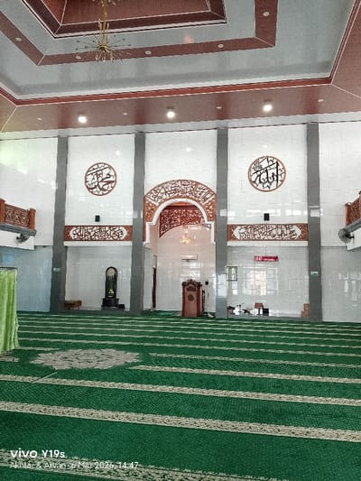 The interior of a mosque is captured in this eye-level, wide-angle shot. The mosque features a spacious prayer hall with a green carpet patterned with rows of white lines, indicating prayer rows. The front wall has three sections separated by grey pillars. Each section is white-tiled and adorned with circular calligraphy art at the top and decorative panels in the middle. The central section has an arched opening leading to another room, also decorated with Arabic calligraphy and featuring a chandelier.

To the left, a green curtain hangs and a small wooden table sits under the balcony. To the right, there is a wall clock with an LED display. The ceiling is multi-tiered with brown and grey panels, and a chandelier hangs from the highest tier.

In the bottom left corner, the watermark "vivo Y19s Akhtar & Arvan 6 Mei 2026, 14:47" is visible. Based on the metadata, the image was taken in Desa Anjatan, Indonesia.