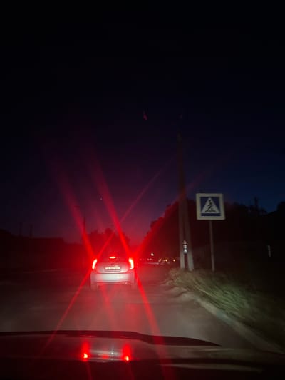 The image depicts a dark, nighttime scene on a road. A white car is visible in the foreground, its red taillights emitting a strong glare. A pedestrian crossing sign with a blue background and a white pictogram of a person walking is mounted on a pole to the right of the road. The road surface appears to be unpaved or poorly maintained. The sky is dark blue, indicating it is likely twilight or night. The scene suggests a rural or semi-urban setting, possibly on the outskirts of Dubăsari, Moldova, given the license plate visible on the white car, which appears to be a Moldovan plate. There are no people visible in the scene.