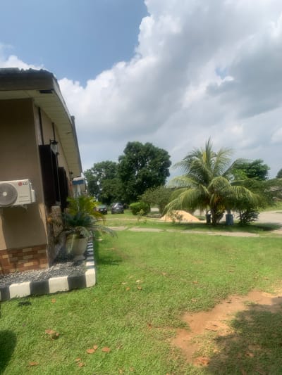 The scene depicts an outdoor area in Akpajo, Nigeria. The left side of the frame shows the exterior of a building with light brown walls and a base featuring a reddish-brown brick pattern. An LG Dual Inverter air conditioning unit is mounted on the wall. Below it, a decorative pot contains a large, leafy plant, possibly a cycad. Two dark, vertical rectangular structures, appearing to be decorative window frames or shutters, are affixed to the wall, each with a light fixture above. The building's base is bordered by a black and white striped curb, which separates a dark gravel bed from a manicured green lawn.

The lawn, which covers the majority of the ground plane, features scattered brown leaves and a reddish-brown dirt patch in the lower right foreground. In the midground, a prominent coconut palm tree stands, bearing coconuts. At its base, two refuse bins, one grey and one blue, are visible. Behind the palm tree, a pile of yellowish material, likely sand or aggregate, is present. Further into the background, a cluster of large, dense green trees is visible. Two dark-colored vehicles are parked on a dirt path or unpaved area between the trees on the left and right sides of the midground. The sky above is bright blue with scattered white cumulus clouds, indicating a partly cloudy day. No individuals are present in the scene, and no specific actions or interactions are depicted.