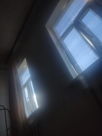 The image depicts an interior scene of a room with two windows. The larger window, located in the upper right, is a skylight type and shows a bright blue sky with horizontal lines, possibly from blinds or textured glass. The smaller window on the left is partially obscured by a light-colored curtain and shows a glimpse of the sky and window frame. The overall lighting suggests it is daytime, with sunlight streaming through the windows, creating bright areas and casting shadows. The environment appears to be a residential interior, possibly a bedroom or living area. There are no people visible in the image. The image lacks specific details to pinpoint its exact location within Aral, Kazakhstan.