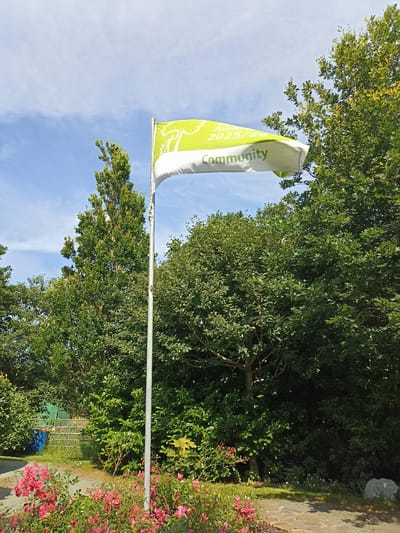 A flag on a flagpole is displayed in front of a dense line of green trees. The flag is light green with white text and graphics. The text reads "Award 2025/26" and "Community". The graphics on the flag appear to depict a stylized tree with figures of people. The sky is blue with some white clouds. In the foreground, there are pink roses and a paved pathway. In the background, there are more trees and some fencing. The scene appears to be outdoors in a park-like setting.
