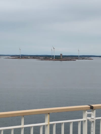 This media file shows a wide shot of a body of water with an island in the distance. On the island, there are several wind turbines and a tall, cylindrical structure that appears to be a lighthouse or tower. The island itself is rocky and sparsely vegetated, with some buildings scattered around. In the foreground, there is a railing, suggesting the image was taken from a boat or ferry. The sky is overcast and a light gray, indicating a cloudy or possibly foggy day. The water is a muted gray-blue, calm and reflecting the sky. The overall impression is one of a somewhat industrial or functional landscape in a maritime setting. There is no visible text in the image.