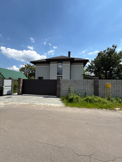 The image depicts a residential street scene in Bucha, Ukraine, under a bright, clear blue sky with scattered white clouds. The setting appears to be a suburban or semi-urban area.
The central subject is a modern, two-story house with light-colored (likely white or light gray) exterior walls and a dark brown or black tiled roof. A dark chimney is visible on the roof. The house features a tall, narrow vertical window on the upper floor and a smaller window on the lower floor.
In front of the house is a property entrance, including a large, dark brown, solid sliding gate. To the right of the gate (from the viewer's perspective), a fence constructed from gray concrete blocks with integrated brick pillars separates the property from the street. A bright yellow gas meter with associated yellow pipes is mounted on this block fence. To the left of the main gate, a smaller metal gate with horizontal corrugated panels and another brick pillar is visible.
An asphalt road, showing signs of cracks, runs along the right side of the image, bordered by a paved sidewalk made of light-colored interlocking pavers. Green grass and weeds grow abundantly along the edges of the road and fence. Behind the main house and fence, a large, leafy green tree provides shade. Part of another house, distinguished by its green roof, is visible in the lower left corner.
The scene is static, with no people or specific activities taking place. The bright lighting suggests it is daytime, likely midday or early afternoon. Text is faintly visible on the yellow gas meter, but it is illegible due to the distance and resolution.