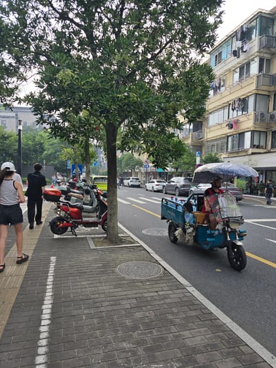 A street scene in Gongshu District, China, features a blue three-wheeled motorized vehicle with an umbrella over the driver, who is wearing a helmet and a red vest. The vehicle carries several bottles and other items. To the left of the road, a sidewalk is lined with trees and parked scooters, including a red one. Two women are walking on the sidewalk, one wearing a white cap and a striped tank top with black shorts, and the other in black pants and a black shirt. Buildings with balconies and laundry hanging out are visible in the background. Cars are driving on the street.