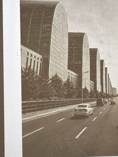 The image depicts a row of monumental, arch-shaped buildings with glass facades, suggesting a modern architectural style. In the foreground, a multi-lane road is visible with several cars driving. Pine trees are planted in front of the buildings. The overall scene appears to be a cityscape under a bright sky.
The architectural style and the specific arrangement of these buildings do not correspond to any known landmarks or significant architectural features in Piacenza, Italy. Therefore, based on the visual evidence in the image, it is not possible to confirm its location within Piacenza.