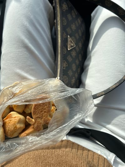 The image shows a partially filled, transparent plastic bag of baked goods, possibly cookies or pastries, held against a backdrop of white and brown fabrics. To the left, a brown Guess brand handbag with a logo pattern and a gold-colored zipper is visible. The lighting suggests the image was taken indoors, with soft natural light illuminating the scene. There is no indication or evidence suggesting the location is Церово, Bulgaria.