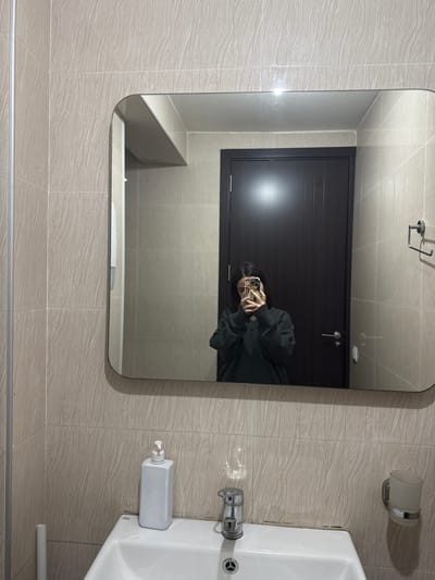 This image depicts an indoor bathroom setting within Bakuriani, Georgia. A person is captured in a mirror selfie, holding a smartphone with a light-colored case to their face, partially obscuring facial features. The individual has dark hair and is wearing a dark, possibly grey or dark green, V-neck long-sleeved top.

The mirror is rectangular with rounded corners and is mounted above a white ceramic sink. On the sink, to the left of a chrome faucet, stands a white, rectangular pump-action soap dispenser. To the right of the sink, mounted on the wall, is a holder containing a frosted glass cup. Above this, a chrome double towel hook is also mounted on the wall. The bathroom walls are covered in light beige or grey rectangular tiles exhibiting a subtle wood-grain or wavy texture. Reflected in the mirror, behind the person, is a dark brown or black paneled door equipped with a silver handle. A vertical silver pipe is visible along the left edge of the image.