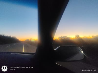 The image captures a car's interior view, looking onto a highway at dawn or dusk. The sky is gradient, transitioning from a light blue to orange at the horizon, where a row of trees is silhouetted. Road markings and the road itself are visible. A car's interior elements, like the dashboard, rearview mirror, and a car pillar, are prominently displayed in the foreground. The photo's timestamp indicates it was taken on November 26, 2025, at 6:39 AM. The image metadata reveals it was shot on a Moto G Stylus 5G in 2024 by Dock Si.