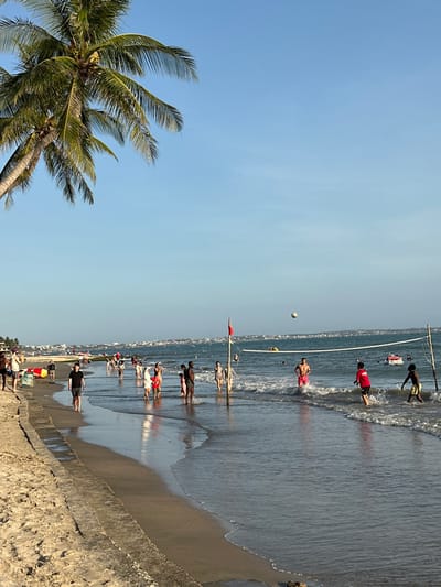 The image depicts a crowded beach scene in Thành phố Phan Thiết, Vietnam, under a clear blue sky. A large palm tree with visible coconuts is partially visible in the upper left foreground. Along the left edge, a concrete retaining wall separates a strip of dry sand from the wet shoreline where gentle waves break. Numerous individuals are present, predominantly in the shallow ocean water.
In the mid-ground, a volleyball net is set up in the water, supported by two posts, one displaying a red flag. Approximately five to eight people are actively playing volleyball in the water; a white volleyball is airborne above the net. Notable players include an individual in a red t-shirt and another in pink shorts.
Further along the shoreline, to the left of the volleyball game, multiple groups of people are wading, walking, or standing in the shallow water. On the immediate left, a person in a black t-shirt and dark shorts walks along the wet sand adjacent to the concrete wall. In the background, various small boats and watercraft, including a yellow inflatable and a white jet ski, are visible on the water. Distant buildings and structures of a coastal town line the horizon.