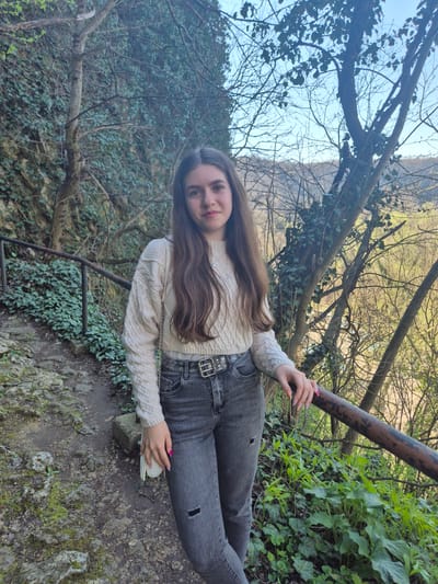 A young woman with long brown hair stands on a rocky path covered in moss and some greenery, next to a weathered metal railing. She wears a beige cable-knit sweater, dark gray jeans with a decorative belt, and holds a small, round white object in her hand. The background features a hillside covered with trees, some bare and some with leaves, and a glimpse of the sky. The location appears to be a natural, wooded area, possibly a park or hiking trail. The photo does not appear to be taken in the city of Ivanovo, Bulgaria, it seems to be taken in a natural environment.
