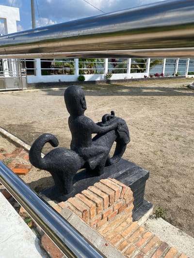 In the city of Parlondut, Indonesia, a black sculpture of a human figure riding a mythical creature is depicted. The sculpture is positioned outdoors on a sandy ground. The creature has a long, curled tail and a circular body, and the human figure is seated atop it, with its arms wrapped around the creature's neck. The sculpture is mounted on a dark base. In the foreground, a section of red brickwork is visible, partially enclosing the base of the sculpture. A metal railing runs across the top of the image, obscuring a portion of the background. The background consists of a paved area, a white building with white fence-like structures, and some vegetation, including palm trees. The sky is blue with scattered clouds.