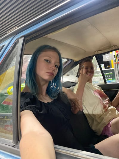 Two young women are seated inside a car, which appears to be at a gas station in Dachniy, Kyrgyzstan. The woman in the foreground, on the left, has blue-dyed hair and is wearing a black off-the-shoulder top. She is looking directly at the camera with a neutral expression. Her right arm is extended towards the camera, and her hand is resting on the car's interior. The second woman, on the right, is wearing glasses and a light-colored t-shirt with a pink graphic. She is sticking her tongue out and making a rock-and-roll hand gesture. The interior of the car is visible, including the ceiling and the seats. Through the car window, elements of a gas station can be seen, including a gas pump with pricing information displayed on a screen. The overall lighting suggests it might be dusk or nighttime.