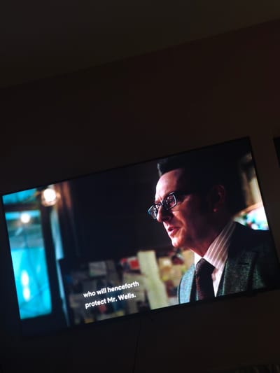 The image shows a television screen displaying a scene with a middle-aged man wearing glasses, a striped shirt, a tie, and a tweed blazer. The text on the screen reads "who will henceforth protect Mr. Wells." The scene appears to be indoors. It is not possible to determine the location in Valchi Dol, Bulgaria, or any specific details about actions, interactions, or environment beyond what is visible on the screen.