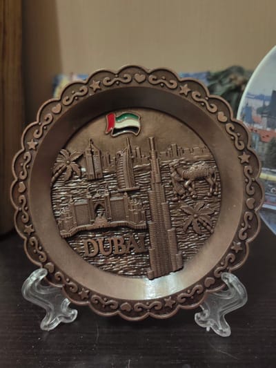 The image shows a close-up, eye-level shot of a bronze-colored decorative plate featuring a relief of Dubai's skyline. The plate has a scalloped edge with a repeating pattern of hearts and swirls, and a star within some of the swirls. At the top center of the plate is a small enamel pin of the United Arab Emirates flag. The relief depicts several skyscrapers, including what appears to be the Burj Khalifa, a palm tree, a camel with a rider, and a building resembling the Burj Al Arab. The word "DUBAI" is embossed in large, blocky letters across the bottom of the relief. The plate is supported by two clear plastic stands. The background is blurred but suggests an indoor setting with wooden furniture and other decorative items. The image does not show the city of Ostafievo, Russia.