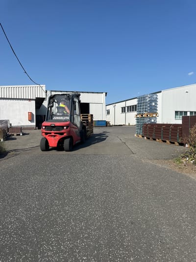 A red Linde forklift, identified by "JUNGBLUH" on its side, is operated by a person wearing a yellow vest and hard hat, driving on an asphalt surface. The forklift is carrying a load of wooden pallets. The scene takes place outdoors, likely in an industrial or commercial area. In the background, there are white industrial buildings with corrugated siding and windows. Several stacks of materials, including what appear to be wrapped metal components and dark brown planks, are visible near the buildings. The sky is clear and blue. The location is identified as Hahn, Germany.