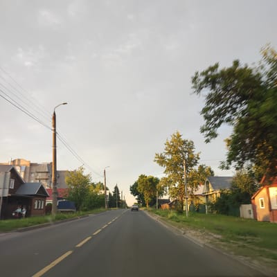 This media file depicts a street scene in Kineshma, Russia, during what appears to be late afternoon or early evening, with the sun casting long shadows. The sky is overcast with soft, diffused light.
The foreground shows a paved road with a yellow dashed line marking the lanes. On the left side of the road, there are several houses and buildings. The closest house is a dark wooden structure with a pitched roof, a porch, and people sitting outside. Further down the street are apartment buildings.
On the right side of the road, there are more residential buildings, including a light-colored house with a grey roof and a brick building with a prominent white window. Green grass and trees line the sides of the road. In the distance, a dark-colored car is driving away from the camera.
A tall, weathered utility pole with multiple wires is prominent on the left side of the road, a common sight in many towns and cities. Another lamppost is visible further down the street. The overall impression is a quiet, residential street in a town, bathed in the gentle light of the setting sun. There is no visible text in the image.