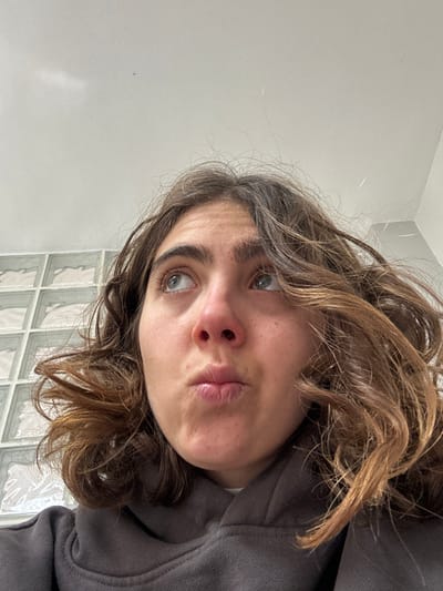 An indoor, low-angle close-up shot depicts an individual with brown, wavy hair and light-colored eyes, looking upwards. The person's lips are slightly pursed. The individual wears a dark-colored hooded sweatshirt. The background includes a plain white ceiling and a section of a wall featuring a grid pattern of translucent, light-colored blocks, likely glass blocks. The specific location within the Menashe Regional Council, Israel, cannot be determined from the provided visual information.