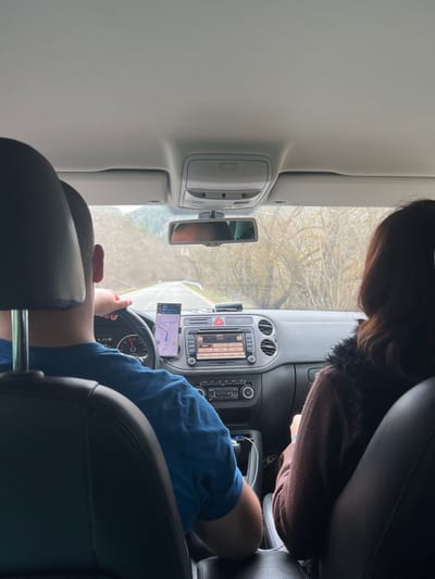 The image depicts the interior of a vehicle from a rear passenger's perspective, looking forward. Two individuals occupy the front seats. A male driver, visible from the back of his head and shoulders, wears a blue shirt and has his hands on the steering wheel. To his right, a female front passenger, also seen from the back of her head and shoulders, has dark brown hair and wears a dark brown garment, possibly with a textured collar.
The dashboard features a smartphone mounted centrally, displaying a navigation application with a map and route. To the right of the smartphone, a black device, possibly a dash camera or radar detector, is affixed to the dashboard. The central console includes a media display unit, air vents, and climate control dials. USB charging cables are visible in the console area between the seats. The rearview mirror reflects the vehicle's headliner and an obscured upper portion of the interior.
Outside the vehicle, a paved road extends straight ahead through an environment characterized by numerous bare, deciduous trees and brush on both sides, indicating a dormant season. Hills or mountains are discernible in the distant background beneath an overcast sky. The vehicle is in motion. This scene takes place in Cherna Mesta, Bulgaria.