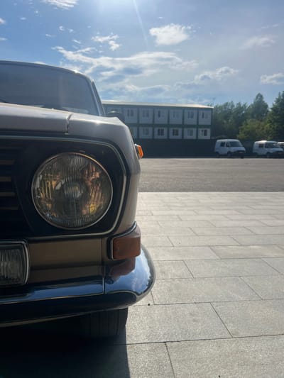 The image shows the front of a classic car, likely a Lada, parked on a paved area. The car is a light brown or beige color with a chrome bumper and a round headlight. An orange turn signal is visible above the headlight. In the background, there are several white modular buildings arranged in rows, possibly temporary housing or storage units. Beyond the buildings, there are trees and a bright blue sky with scattered white clouds, suggesting it is a sunny day. Two white vans are parked in the distance. The sun is low in the sky, casting long shadows and creating a lens flare on the left side of the image, indicating it is either early morning or late afternoon. The scene appears to be an outdoor parking lot or service area. There is no visible text in the image.