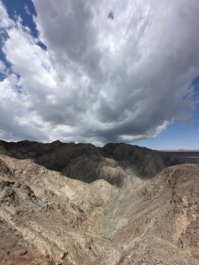 The image displays a panoramic view of a rugged, mountainous desert landscape under a dramatic sky filled with heavy, gray clouds interspersed with patches of blue. The foreground features steep, rocky slopes with varied textures and colors, ranging from light tan and gray to reddish-brown, indicative of exposed rock and sparse vegetation. The mid-ground is dominated by dark, craggy mountain peaks that recede into the distance, their surfaces deeply eroded and casting long shadows. A dry, barren valley can be seen nestled between some of the mountains. In the far background, a flat, desolate plain stretches towards the horizon, where it meets the sky. The overall impression is one of a remote and arid environment.
This scenery is characteristic of the region surrounding Jiayuguan, China, particularly the foothills of the Qilian Mountains, which lie to the south of the city. The area is known for its harsh, desert climate and dramatic geological formations. While the specific location within or near Jiayuguan is not precisely identified in the image, the landscape is consistent with areas like the Gobi Desert or the mountainous terrain that forms the natural defenses of the Jiayu Pass. No people or human structures are visible in the immediate scene. The weather appears overcast and potentially unsettled, with the dark clouds suggesting possible rainfall or a storm approaching.