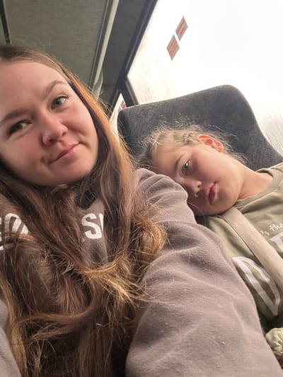 The image depicts a close-up, upward-angled selfie of two females in an indoor setting, identified as Kvarsa, Russia, within a mode of transport such as a bus or train. In the left foreground, a young adult female with long brown hair, light-colored eyes, and a faint smile is partially visible, wearing a light brown or taupe hooded sweatshirt with white lettering, of which "S" is clearly discernable. Her arm, clad in the sweatshirt, extends across the lower portion of the frame. To the right and slightly behind her, a child, likely pre-teen, with light hair styled in small braids, rests her head on a dark grey fabric seat. The child's eyes are open, looking towards the camera, and she wears an olive green long-sleeved shirt with partially visible white lettering ("s the"). A light-colored seatbelt is fastened across her chest. In the background, a bright, overexposed window illuminates the scene, and two small red-orange rectangular labels or signs are affixed to an overhead panel or window frame. A grey ceiling or overhead panel with subtle linear details is visible above the young adult female's head.