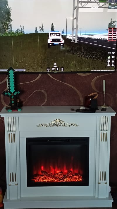 A screen displays a driving simulator game, showing a white SUV with flashing blue lights on a grassy field next to a highway. A digital display indicates "0 KM/H" and "A/T 1". Below the screen, a white electric fireplace with faux glowing logs is visible. On the mantlepiece above the fireplace, to the left, is a teal pixelated sword decoration and a glass bottle. To the right, there are a few electronic devices, a white candle in a gold holder, and a dark red lantern. The background is a patterned wallpaper. The scene is indoors, likely in a residential setting. No people are present. The context of Volgodonsk, Russia, is not discernible from the image itself.