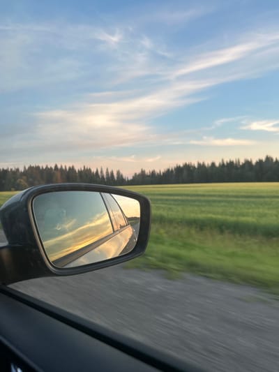 The image shows a view from inside a car, looking out the side window and into the car's side mirror. The side mirror reflects a sunset sky with wispy clouds and the orange and yellow hues of the setting sun. Outside the car, a blurred green field stretches towards a dark line of trees that forms the horizon. The sky above is a clear blue with scattered white clouds. The road, also blurred due to motion, is visible in the foreground and the bottom of the frame. The car appears to be driving through a rural area, likely in the city of Glazunovo, Russia, given the landscape. There are no people visible in the scene.