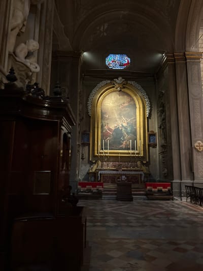 This image depicts the interior of the Chiesa di San Martino (Church of Saint Martin) in Ferrara, Italy, viewed from a nave or side aisle looking towards the main altar.
The central focal point is a large, ornate altarpiece featuring an arched-top oil painting, *The Vision of Saint Bruno* by Guercino, set within a prominent golden frame. The painting shows multiple figures, including angels and a saint, illuminated by a light source depicted in the upper left. Below the painting, a marble altar table is covered with a white cloth over a dark red frontal. Four tall, golden candlesticks with white candles and one shorter candlestick are arranged on the altar table. A dark wooden lectern or stand is positioned directly in front of the altar table.
The church interior is characterized by tall, dark-toned walls adorned with pilasters and moldings. Above the altarpiece, an octagonal stained-glass window displaying blues, reds, and yellows is set high in the arched wall. Protective netting is visible across the vaulted ceiling above. On either side of the altarpiece, vertical niches house statuary. Gold trim highlights architectural elements and frames. To the far right, a four-lobed decorative element is visible on the wall.
In the left foreground, a dark wooden confessional booth or large pew partially obscures the view, featuring carved details and a rectangular panel. Above and to the left of this structure, a white marble sculpture of a cherub or putto is mounted on the wall. The floor is covered with a geometric pattern of light and dark tiles. On the right side of the image, several dark wooden chairs are arranged along a wall, and two lower, red-draped wooden structures flank the main altar. The scene is dimly lit, with specific illumination on the altarpiece and the stained-glass window. No individuals are present.
