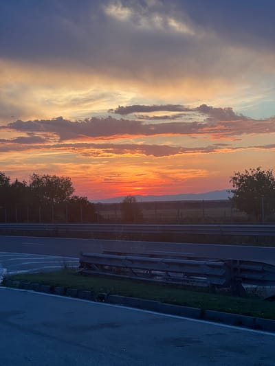 The image captures a highway scene at dusk, with a vibrant sunset painting the sky with hues of orange, yellow, and purple. A barrier separates the road from a grassy area and a treeline. In the distance, a flat landscape is visible, possibly with mountains on the horizon. The road surface is dark, suggesting it might be asphalt. The time of day and the presence of a highway indicate the location is likely near a transit route. While the specific location is identified as Orizovo, Bulgaria, no inhabitants or specific actions are discernible in the image.