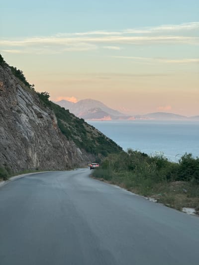 An asphalt road curves to the right, flanked by a steep, rocky mountainside on the left and a body of water and distant mountains on the right. A light-colored car with its brake lights illuminated is driving on the road in the distance. The sky transitions from a pale blue at the top to a soft pink and orange hue near the horizon, with thin clouds stretching across it. The landscape is predominantly green with dense vegetation growing on the slopes. The scene appears to be captured during either sunrise or sunset, with the lighting suggesting a temperate climate. The location is the city of Višnjevo, Montenegro.