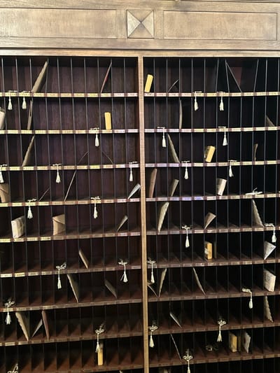 The image captures a large, vintage wooden pigeonhole rack, likely a hotel key and message organizer, set indoors. The rack is made of dark stained wood and features numerous square and rectangular compartments. To the left, a section of lighter wooden paneling with a carved geometric design is visible.
Many compartments have small, light-colored rectangular labels bearing numbers. Visible text includes "105", "106", "107", "108", "109", "110", "111", "112", "227", "233", "234", "235", "323", "324", "329", "407", "408", "409", and "520-", indicating room numbers or mailbox identifiers.
Within the compartments, various items are stored. These include numerous folded envelopes or pieces of paper, likely mail or messages. Many compartments also feature small metal hooks with white, tasseled key fobs or room key accessories hanging from them. Some slots contain small, plain rectangular objects, possibly room keys.
No people are present, and no active event is taking place; the system appears to be a static storage and organization unit. The setting is indoors, with artificial lighting. The style of the pigeonhole rack suggests an older, traditional establishment, possibly a historic hotel or inn in the La Quinta area of the United States. The overall impression is one of organized, functional nostalgia.