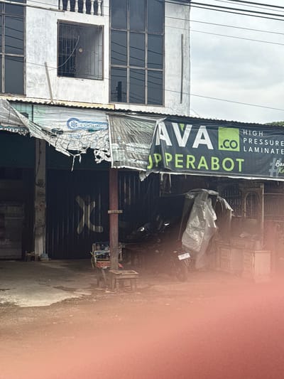 The image depicts an outdoor scene in front of a commercial building, likely in Lamreung, Indonesia, given the context provided. The building has a white, weathered facade with multiple windows. A large, tattered banner stretches across the front, advertising "AVA .CO HIGH PRESSURE LAMINATED" and "UPPERABOT". Beneath the banner is a dark, corrugated metal storefront. A motorcycle, partially covered with a light-colored tarp, is parked in front of the storefront. Various items, including what appear to be bottles and a cart, are also visible near the entrance. The ground is paved, and there is evidence of dust or dirt. The overall impression is one of a somewhat dilapidated, functional business establishment in an urban or semi-urban setting. There are no people clearly visible in the image.