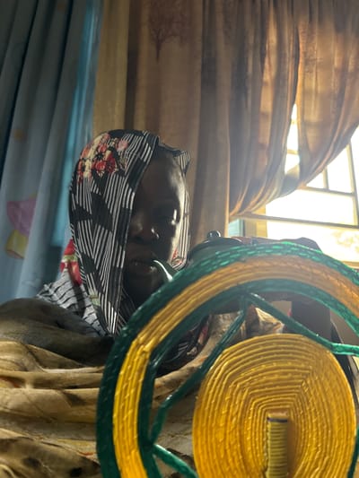 A person with a headscarf, lying down on a blue surface with a yellow and green woven circular object. The person is likely indoors in a dwelling with window curtains. The location is Madangala, Nigeria.