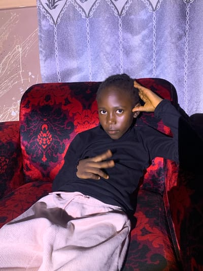 A young Black person is sitting on a red and black patterned sofa. They are wearing a black turtleneck shirt and light pink pants. Their left hand is resting on their head, and their right hand is held up in front of them, with fingers splayed. The background consists of a wall with white splatters and a purple curtain with a decorative pattern. The lighting suggests an indoor setting, possibly a home in Russo, Nigeria.