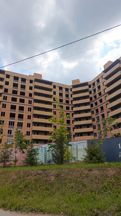 The image shows a large, multi-story building under construction in Zvenigorod, Russia. The building is made of brick and has many windows and balconies. There are trees and grass in front of the building, and a billboard with an image of a completed building and text is visible. The sky is cloudy.
