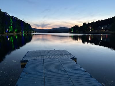 Captured at dusk in Dom Otdykha Zolotoy Plyazh, Russia, the image features a serene lake reflecting the twilight sky and the surrounding landscape. A dark blue floating dock extends from the bottom center of the frame, creating a sense of depth and leading the eye into the scene. On the left and right shores, lush trees line the water's edge. The trees on the left are illuminated with blue and green lights. On the right side, buildings are visible amongst the trees with lights shining. In the distance, silhouettes of mountains can be seen beneath the colorful sky, adding to the picturesque and tranquil atmosphere. The lake's surface is mostly still, enhancing the mirror-like reflection.
