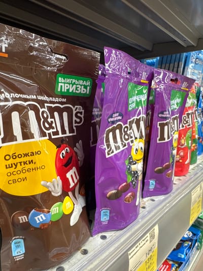 This media file displays a close-up, slightly angled shot of several bags of M&M's candy arranged on a store shelf. The dominant colors are purple, brown, and yellow from the candy packaging, with some blue and green also visible. The products appear to be M&M's candies, with some bags featuring purple packaging and depictions of M&M characters and blueberries, suggesting a "blackcurrant" flavor. Another bag in the foreground is brown and features the M&M's logo and characters, along with Russian text.
Visible text on the packaging includes "ВЫИГРЫВАЙ ПРИЗЫ" (Win Prizes), "молочным шоколадом" (with milk chocolate), and nutritional information like "ккал 191" (kcal 191) and "8%". Other text visible on the purple bags includes "80г" (80g) and descriptions possibly related to the flavor. There are also price tags visible on a shelf above the candy, showing prices like "139" and "147".
The setting is clearly a retail environment, likely a supermarket or convenience store, indicated by the shelves and the organized display of products. The lighting is bright, typical of indoor retail lighting, and there are no discernible environmental cues related to time of day or weather. The focus is entirely on the candy products, with no people or animals present. The overall impression is one of commercial display, highlighting the product's branding and potential promotions.