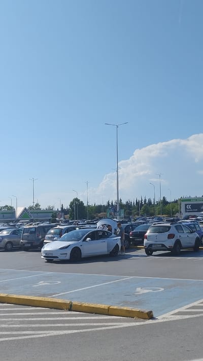 A wide-angle, full-shot photograph captures a crowded outdoor parking lot on a clear, sunny day in Pylaia Municipal Unit, Greece. The foreground shows a section of the asphalt parking lot marked with blue and white lines, including a blue-painted space designated for disabled parking with a white wheelchair symbol. A low, yellow curb runs along the edge of the parking bay.

In the middle ground, numerous cars are parked in rows. A prominent white Tesla Model 3 is parked in the foreground, facing left, with its rear trunk open. Three individuals are visible near the Tesla; two men and one woman. One man, wearing a grey t-shirt and shorts, stands beside the open trunk. Another man, wearing a white t-shirt, is partially visible behind the Tesla. A woman, wearing a white top and dark shorts, is also near the open trunk. Other vehicles in the middle ground include a white Dacia Sandero, a silver hatchback, and a dark-colored SUV.

In the background, several tall streetlights are visible. Beyond the parking lot, a building with a green and white sign that reads "LEROY MERLIN" is present. Further in the distance, a line of trees and a slightly hilly landscape are visible under a bright blue sky with scattered white clouds. The overall scene suggests a busy commercial area, likely a shopping center or retail park.