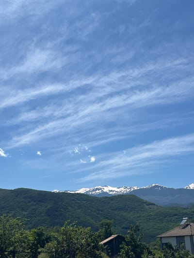 The image shows a landscape scene in Bukovo, Bulgaria. The sky is predominantly blue with streaks of wispy white clouds running horizontally across the frame. A mountain range is visible on the right side, with snow-capped peaks near the top. Lush green trees cover the lower slopes of the mountains. Parts of buildings are visible on the upper right-hand side, including a roof and solar panels. Trees obscure the view towards the bottom right corner.